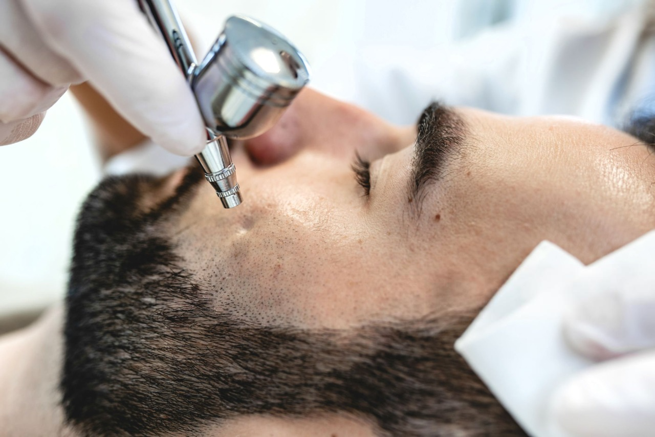 A person receives a facial treatment with a spray device while lying down, as a gloved hand holds a cloth to their face.