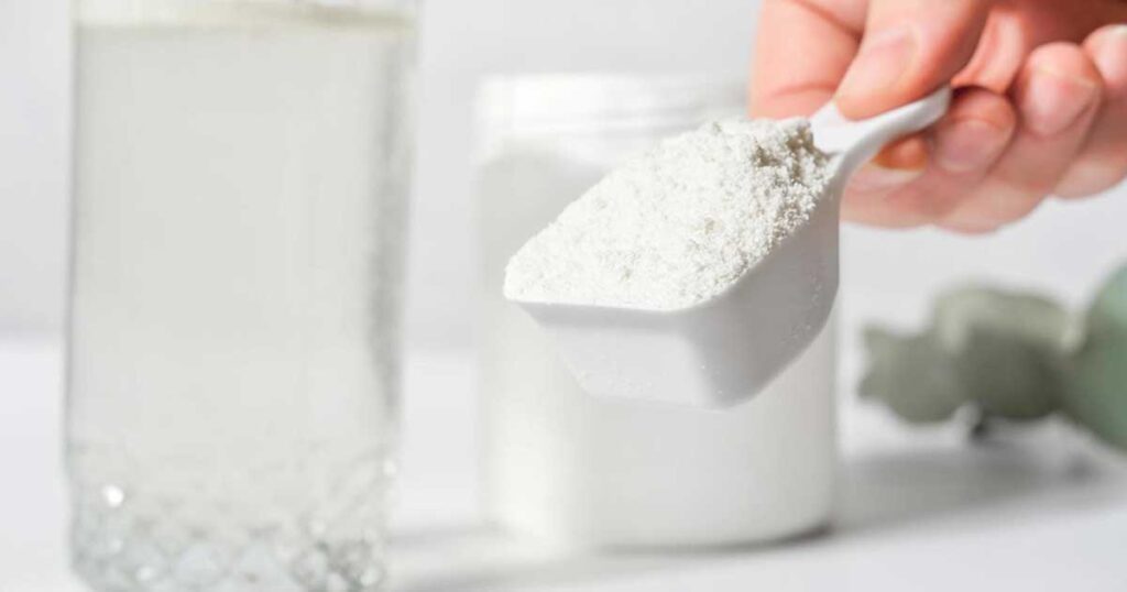 A hand holding a scoop filled with collagen peptides next to a glass of water